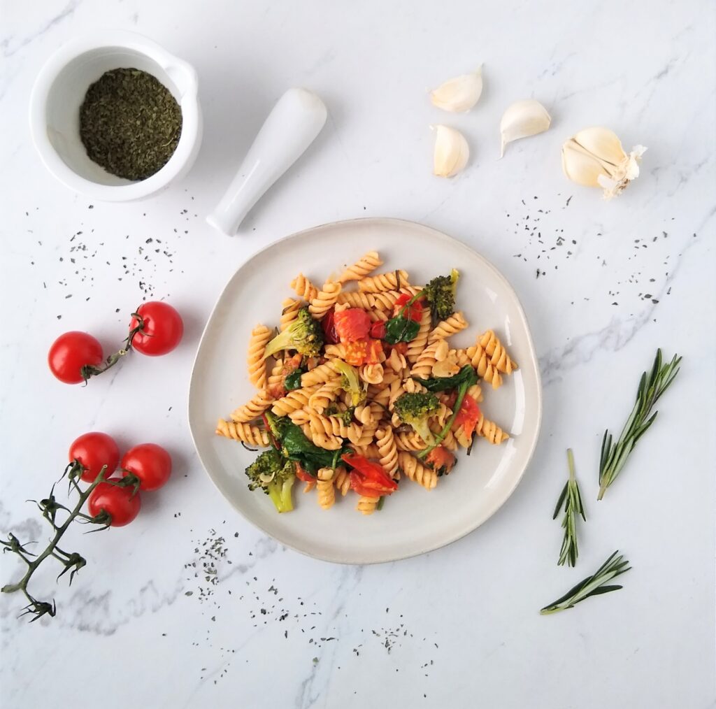 a dish of pasta with tomatoes and broccoli, surrounded by a vine of cherry tomatoes, herbs, garlic cloves, and a mortar and pestle with herbs