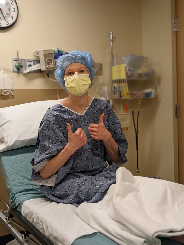 woman with a mask and hairnet, sitting on a hospital bed with two thumbs up