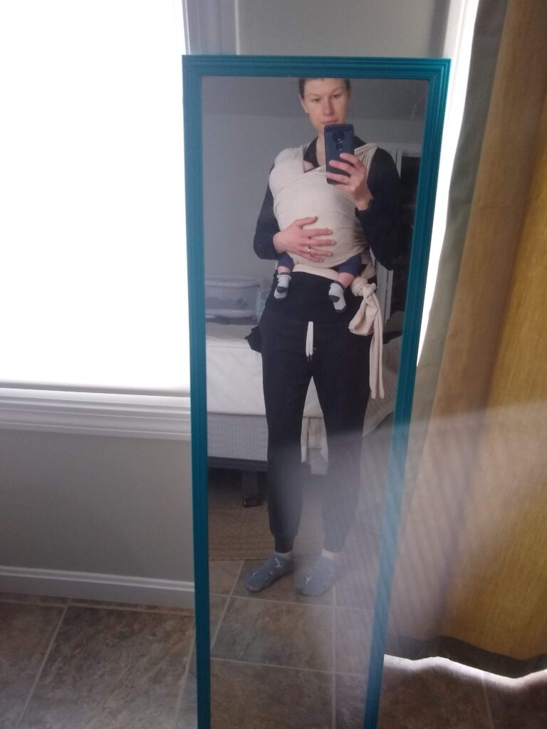 woman wearing sweats standing in front of a mirror, wearing a baby in a wrap