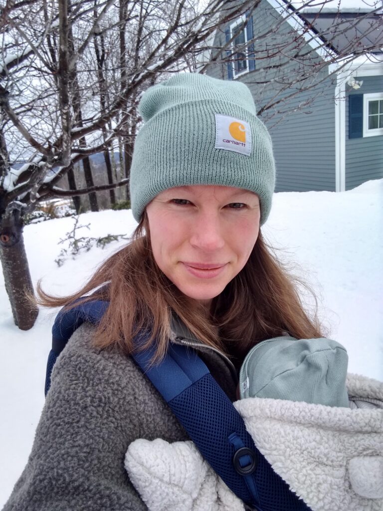 woman outside, snow is on the ground, with a winter hat on and a baby bundle up in a carrier