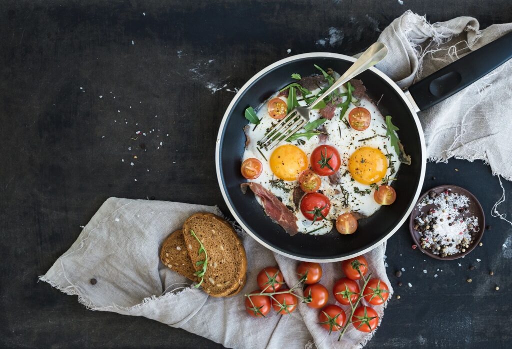 skillet with fried eggs and whole cherry tomatoes, some arugula leaves. It's set on a table with cloth, a small dish of salt, a small vine of cherry tomatoes, and two small slices of bread