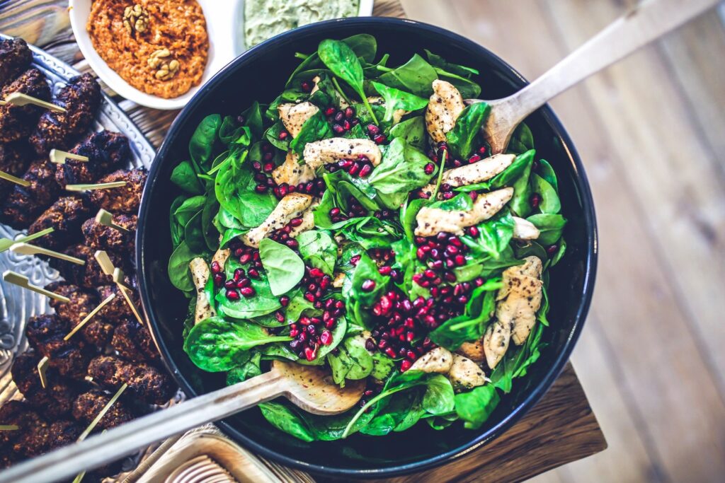 a bowl of spinach topped with chicken and pomegranate seeds and salad serving utensils sticking out