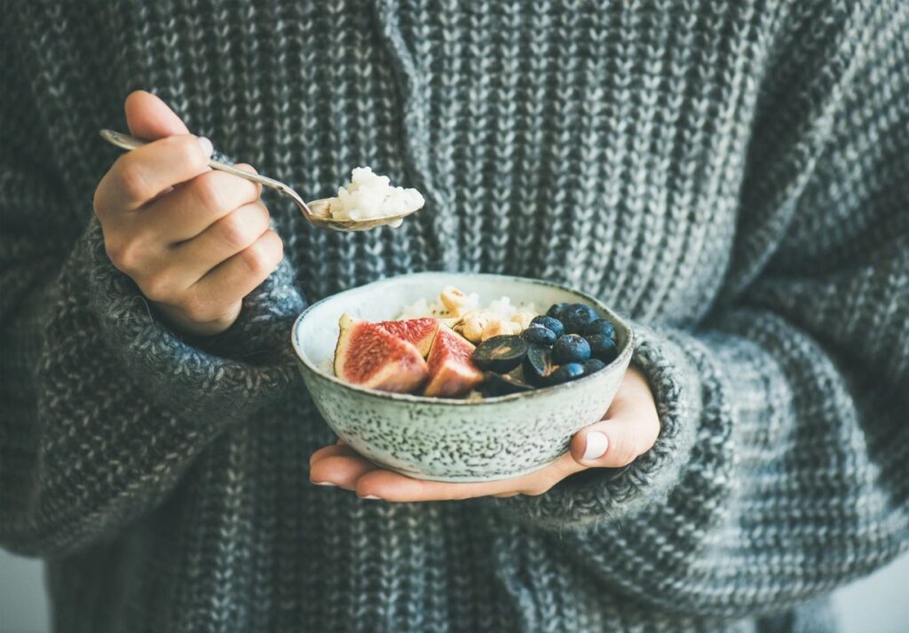 person in a sweater holding a bowl of oatmeal and fruit